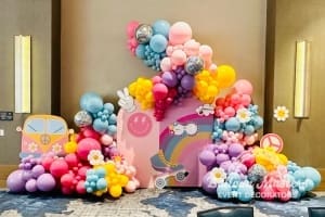 Purple, pink, blue, and yellow balloon display with balloon flowers, disco balls and 60s cut outs scattered in balloons.