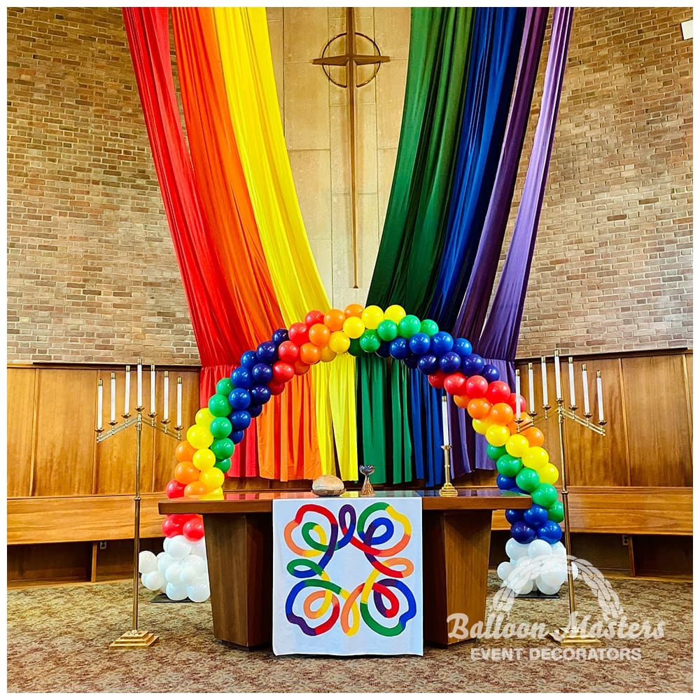 Pride rainbow balloon display with draped rainbow fabrics behind the altar of a church.