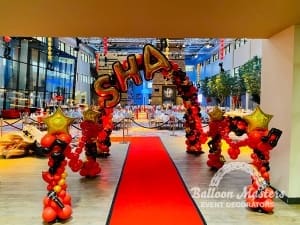 A red, black and gold balloon arch above a red carpet with text "SHA" at the top. There are black and red balloon guardrails along sides of red carpet.