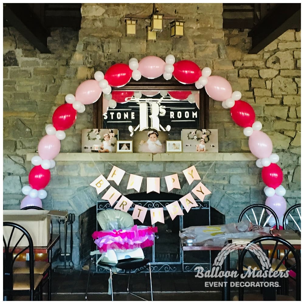 A pink, red and white balloon arch over stone fireplace.