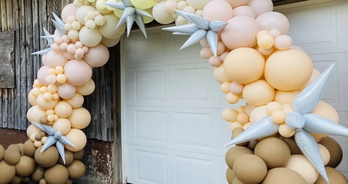 Brown, orange, peach, and yellow balloon arch with silver stars attached infront of garage.