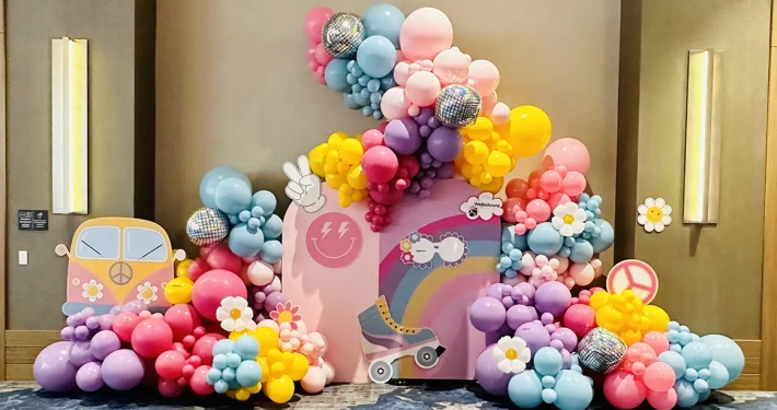 Purple, pink, blue, and yellow balloon display with balloon flowers, disco balls and 60s cut outs scattered in balloons.